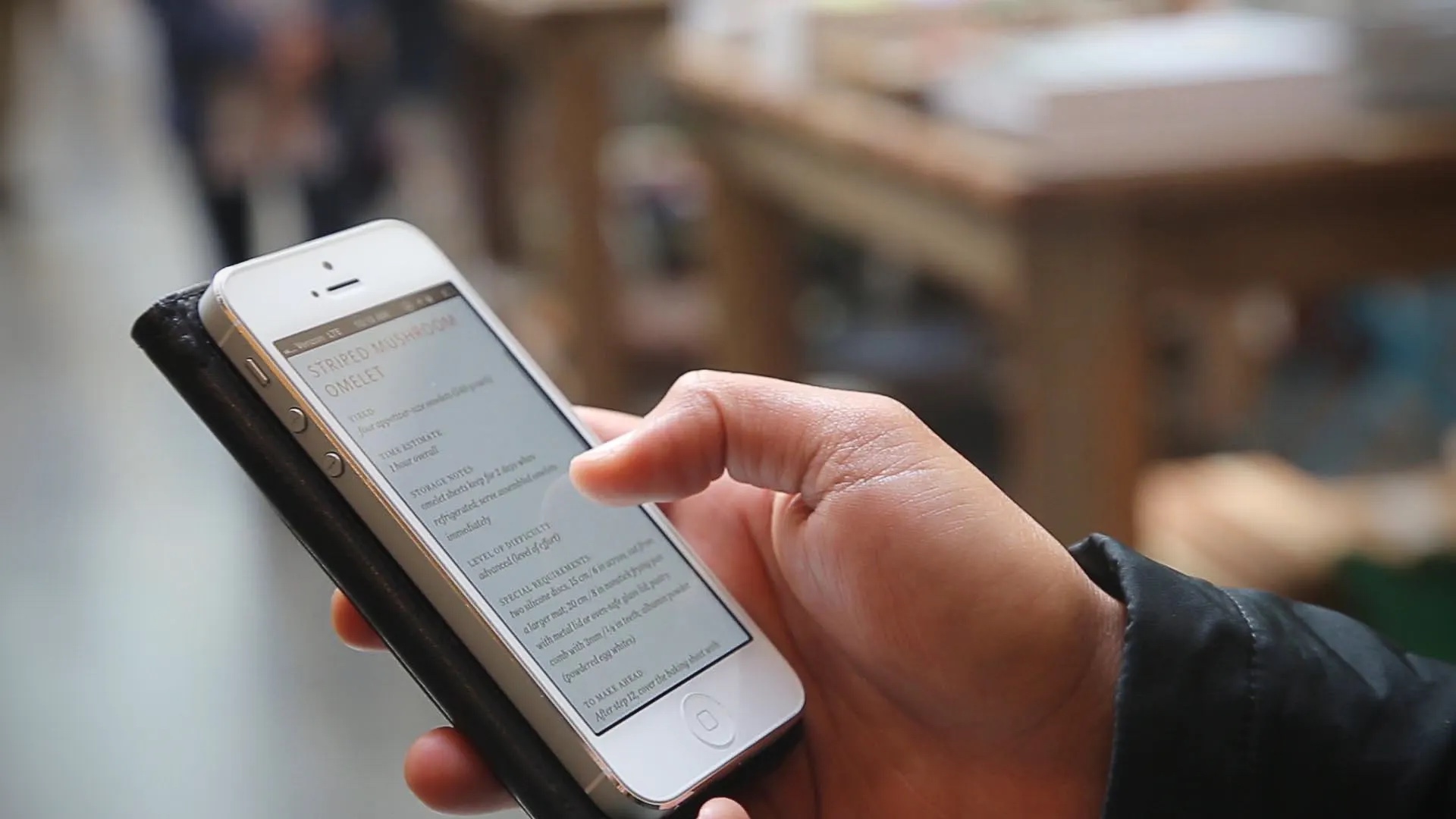 A thumb scrolling a recipe in Inkling on an iPhone 5 held in a black case. The screen shows a striped mushroom omelet recipe.