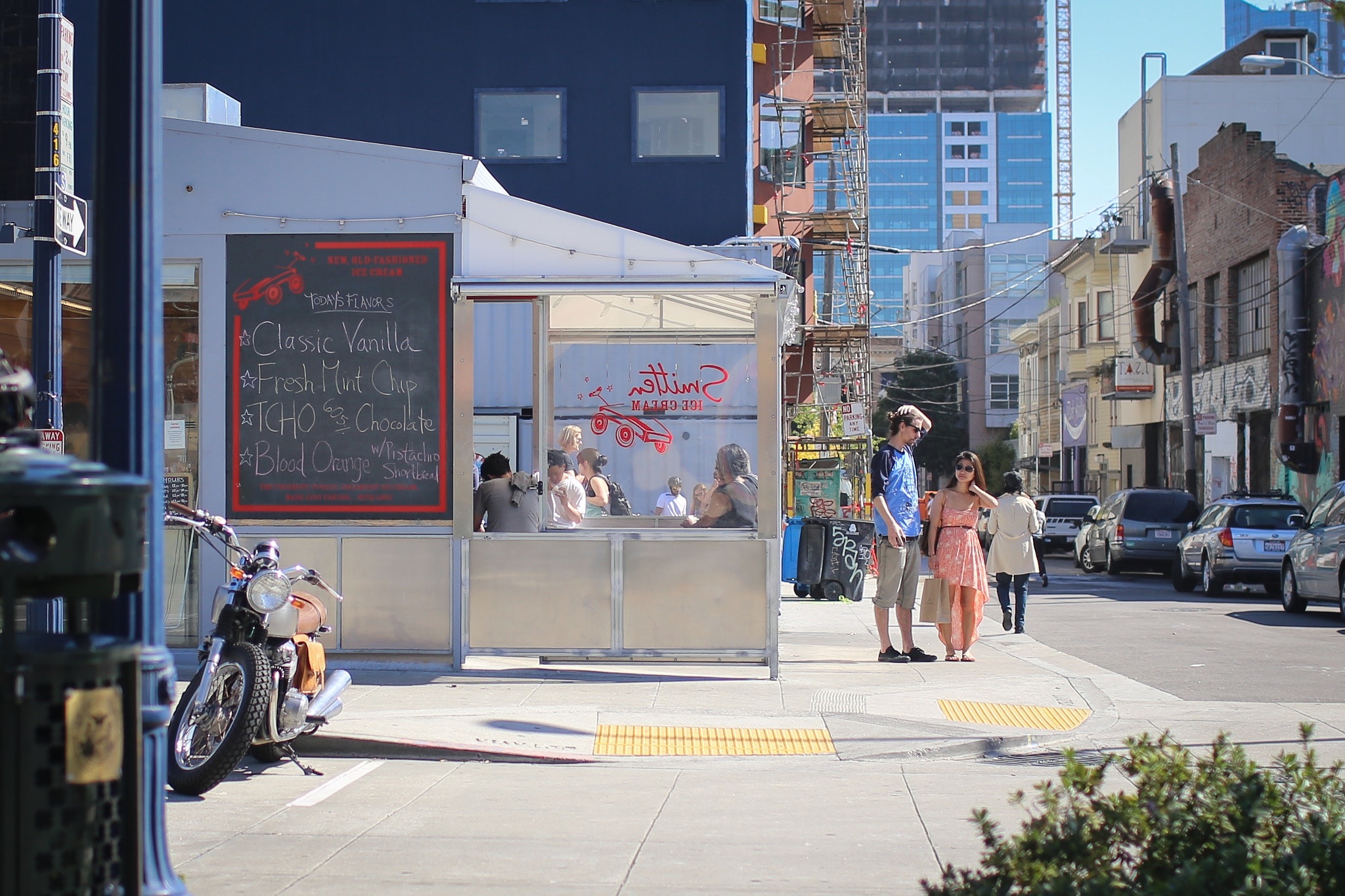 An ice cream stand on a San Francisco street corner, with a chalkboard menu, a parked motorcycle, and people in line.